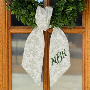 Puede incluir: Una cinta blanca con un estampado floral verde y las iniciales verdes bordadas "MBR" cuelga de una corona verde. La cinta está atada con un lazo y es visible contra una puerta de madera.