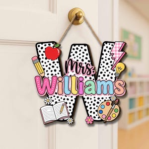 May include: A wooden door sign in the shape of the letter "W" with a black and white polka dot pattern. The sign says "Mrs. Williams" in colorful letters and features school-themed decorations like an apple, pencil, and paint palette.