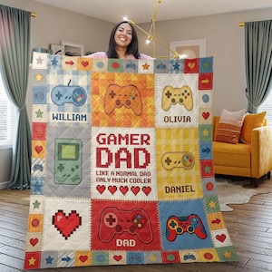 May include: A personalized gamer-themed blanket with a patchwork design. It features pixelated hearts, game controllers, and the text "Gamer Dad" with names like William, Olivia, and Daniel. The blanket has a variety of colors including blue, red, and yellow.