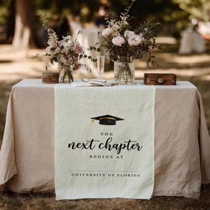 May include: A graduation-themed table runner with the text "The Next Chapter Begins At University of Florida" and a mortarboard graphic. The runner is cream-colored and placed on a table with floral arrangements.