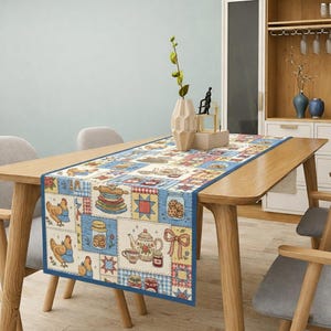 May include: A rectangular table runner with a patchwork design featuring kitchen-themed illustrations. The runner has a blue border and showcases images of chickens, jars of cookies, teapots, and rolling pins. The runner is placed on a wooden table.