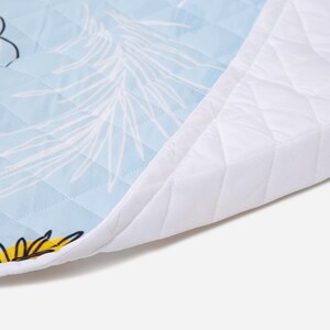 May include: A quilted fabric mat featuring a light blue background with white line-drawn palm leaf patterns and yellow floral accents. The mat has a solid white quilted border and a diamond-stitched texture throughout the entire surface.