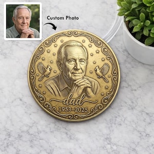 May include: A round, bronze-colored memorial coin featuring a portrait of a man with the word "dad" and the years 1953-2025. The coin is decorated with stars and bird illustrations. A small photo of the man is in the upper left corner.