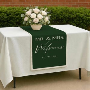 May include: A white tablecloth with a dark green table runner featuring the text "MR. & MRS. Wilsons" and the date "01.03.26". A vase of white roses sits on the table. The table is outdoors.