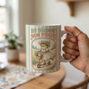 May include: A ceramic mug with a whimsical design. The mug features a green and white striped background and the text "OFF TO COMMIT MOM FOOLERY". An illustration of a frog riding a scooter is in the center. The frog is wearing a dress and hat.