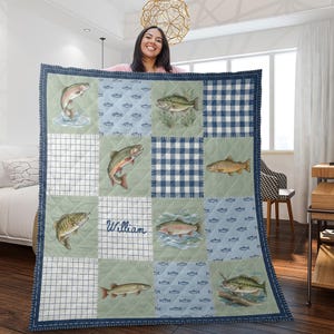 Peut inclure: Une couverture matelassée avec un motif patchwork composé d'illustrations de poissons et de carreaux bleus et blancs. La couverture a une bordure bleu marine et le nom "William" est écrit sur l'un des carrés.