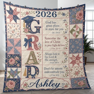 May include: A quilted graduation blanket with the year 2026 and the name "Ashley". The design includes the word "GRAD" in large letters, floral patterns, and inspirational quotes. The color scheme features pink, blue, and cream.