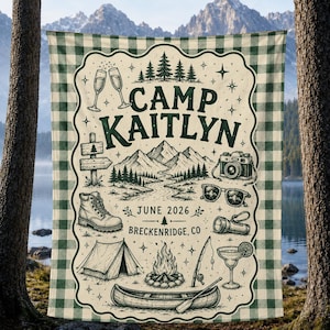 May include: A rectangular blanket with a green and white buffalo plaid border. The center features a cream background with dark green illustrations of mountains, a tent, a canoe, a campfire, a hiking boot, a camera, sunglasses, and text reading CAMP KAITLYN JUNE 2026 BRECKENRIDGE, CO.