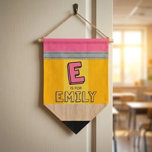 May include: A fabric wall banner shaped like a pencil, featuring a pink eraser top, a grey metal band, a yellow body, and a black pointed tip. The banner displays the letter E, the text IS FOR, and the name EMILY in black outlined lettering.