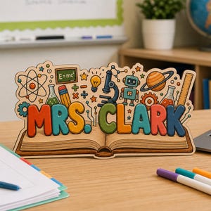 May include: A wooden desk sign featuring the name MRS. CLARK in colorful 3D-style lettering, resting on an open book illustration. The design includes icons of a microscope, robot, planet, lightbulb, and a chalkboard with E=mc2 written on it.
