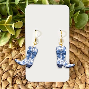 May include: A pair of earrings featuring a cowboy boot design. The boots are blue and white with a floral pattern and a blue bow. The earrings have gold-colored hooks and are displayed on a white card.