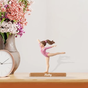 May include: A figurine of a gymnast in a pink leotard, balancing on one leg with arms outstretched. The figurine is on a wooden base with the name "Hailey" engraved. A vase of flowers and a clock are in the background.
