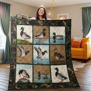 Könnte beinhalten: Eine gesteppte Decke mit neun quadratischen Feldern, die jeweils ein anderes Bild einer Stockente zeigen. Die Enten werden in verschiedenen Posen vor Hintergründen aus Wasser, Gras und Erdtönen dargestellt. Der Rand hat ein Tarnmuster.