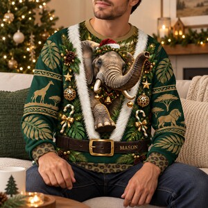 May include: A dark green long-sleeve sweatshirt featuring a central 3D-style elephant wearing a Santa hat, surrounded by white faux fur trim, candy canes, and ornaments. The waist area displays a brown belt graphic with the name MASON printed on it.