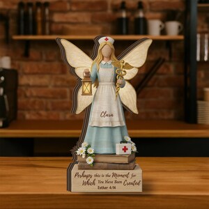 Pode incluir: Estatueta de anjo de madeira com uniforme de enfermeira, segurando uma lanterna e um símbolo médico. A estatueta tem cabelo loiro e asas. A base tem o texto "Perhaps this is the Moment for Which You Have Been Created Esther 4:14".