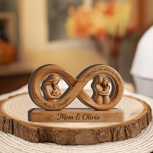Puede incluir: Un adorno de madera con forma de infinito que presenta dos recortes con figuras talladas de una madre sosteniendo a un bebé y una madre abrazando a su hija. La pieza está grabada con el texto "Mom & Daughter", "A bond that can never be broken" y "Mom & Olivia".