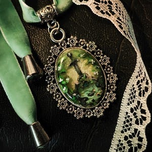 May include: A silver pendant necklace with a green and white resin cameo featuring a silver anchor. The pendant is surrounded by a decorative silver frame with a floral design. The necklace is on a green velvet ribbon.