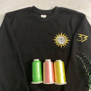 May include: Black sweatshirt with gold embroidered sun and face design, and the number 33 on the sleeve. Three spools of thread in green, pink, and yellow are in front of the sweatshirt.