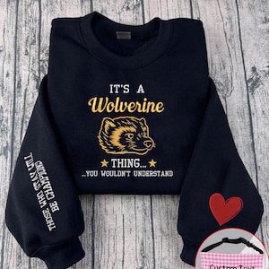 May include: A navy blue sweatshirt featuring the text "IT'S A Wolverine THING... YOU WOULDN'T UNDERSTAND" in gold. The sleeve displays the text "THOSE WHO STAY WILL BE CHAMPIONS". A red heart and a pink gingham pet bandana are also visible.