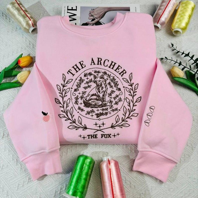 K&ouml;nnte beinhalten: Rosafarbener Pullover mit einem braunen gestickten Design, das einen Fuchs in einem Kreis mit dem Text "THE ARCHER" und "THE FOX" darunter zeigt.
