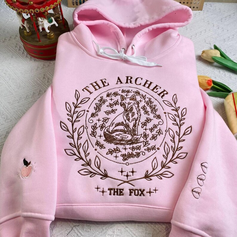 K&ouml;nnte beinhalten: Ein rosa Hoodie mit einem braunen Stickmuster, das einen Fuchs, Bl&auml;tter und den Text "THE ARCHER" und "THE FOX" zeigt.