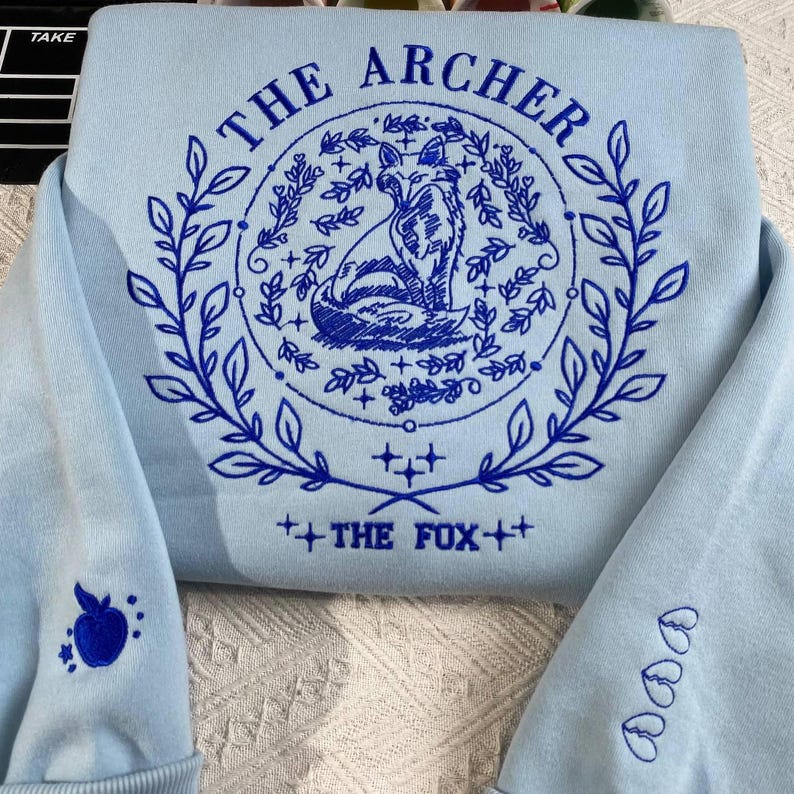 K&ouml;nnte beinhalten: Hellblauer Pullover mit blauer Stickerei, die einen Fuchs, Bl&auml;tter und den Text "The Archer" und "The Fox" zeigt.