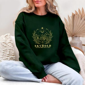 May include: Dark green sweatshirt with a gold graphic of a castle, mountains, and celestial elements. The word "SKYHOLD" is printed below the graphic, with the phrase "THE PLACE WHERE THE SKY IS KEPT" underneath. The sweatshirt is a solid color.