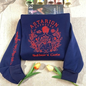 May include: A navy blue sweatshirt with a red embroidered design featuring a figure with a crown of thorns, surrounded by clouds, moons, and suns. The text "Astarion Approves" and "Baldur's Gate" are also embroidered in red.