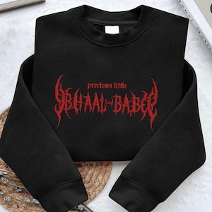 Könnte beinhalten: Schwarzes Sweatshirt mit dem roten Schriftzug "precious little BHAAL BABE" in einer stilisierten gotischen Schriftart. Das Kleidungsstück ist gefaltet, die Ärmel sind gekreuzt. Der Text ist mittig auf der Brust platziert.