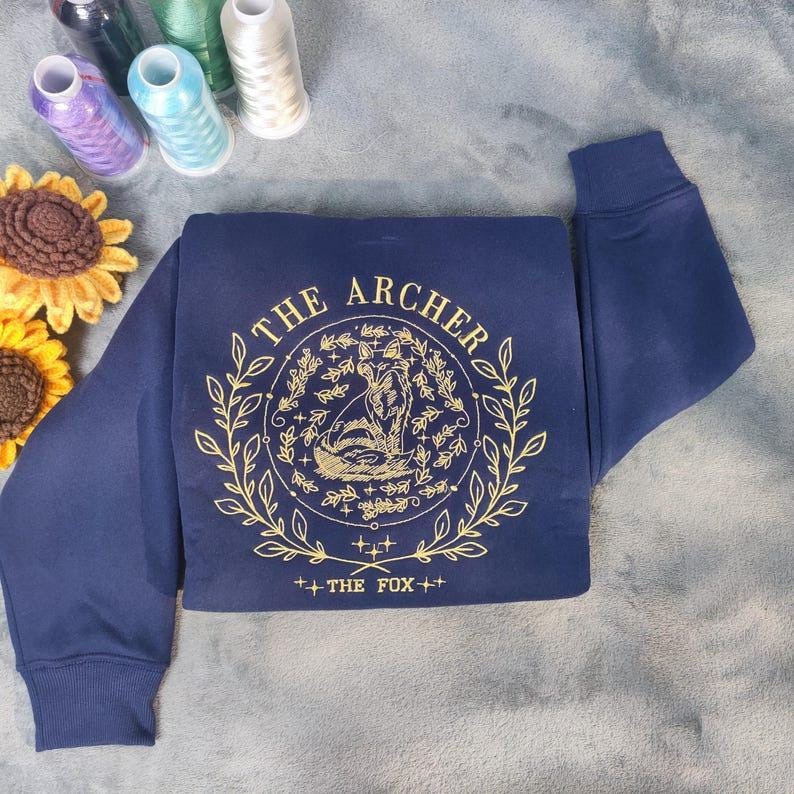 K&ouml;nnte beinhalten: Marineblauer Sweatshirt mit einem goldenen Stickmuster, das einen Fuchs in einem Kreis mit dem Text "THE ARCHER" und "THE FOX" darunter zeigt.