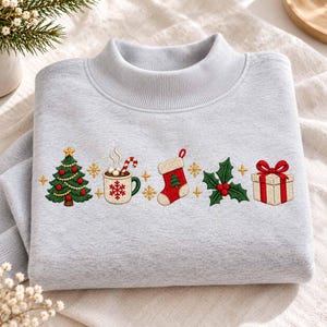 May include: A light grey mock neck sweatshirt featuring embroidered Christmas icons including a decorated green tree, a mug of cocoa with a candy cane, a red stocking, holly leaves with red berries, and a gift box with a red bow.