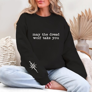May include: Black sweatshirt with the text "may the dread wolf take you" in white. A white graphic of a sword and sunburst design is on the left sleeve. The sweatshirt is a casual style.