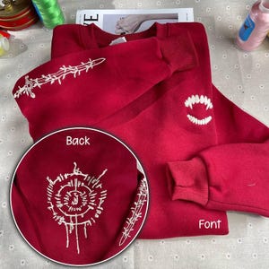May include: Red sweatshirt with white embroidered designs. The back features a circular design with a sword and the front features a sword and fangs.