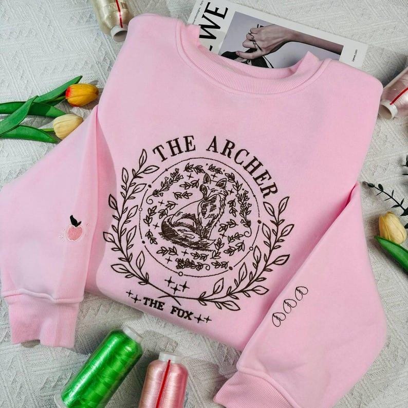 K&ouml;nnte beinhalten: Rosafarbener Pullover mit einem braunen gestickten Design, das einen Fuchs in einem Kreis mit dem Text "THE ARCHER" und "THE FOX" darunter zeigt. Der Pullover hat ein kleines gesticktes Apfelchen am linken &Auml;rmel.