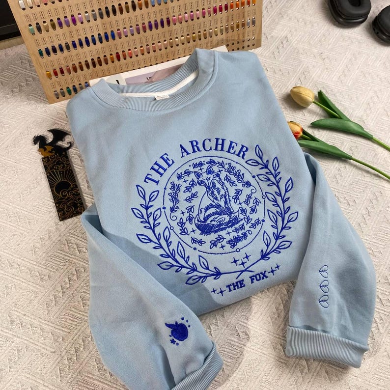 K&ouml;nnte beinhalten: Hellblauer Pullover mit einem blauen gestickten Design eines Fuchses in einem Kranz mit dem Text "The Archer" und "The Fox" auf wei&szlig;em Hintergrund.