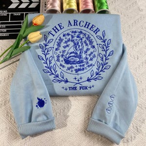 May include: A light blue sweatshirt with a blue embroidered design featuring a fox in a circle with the text "THE ARCHER" and "THE FOX" on a light blue background.