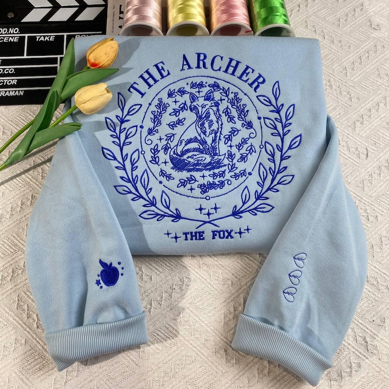 K&ouml;nnte beinhalten: Ein hellblauer Pullover mit einem blauen Stickmuster, das einen Fuchs in einem Kreis mit dem Text "THE ARCHER" und "THE FOX" auf einem hellblauen Hintergrund zeigt.