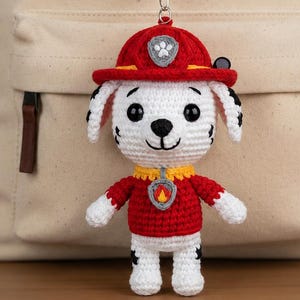 May include: A crochet amigurumi dog keychain wearing a red firefighter hat and uniform, featuring black eyes and a fire badge. The text reads Mini Rescue Puppy Crochet Pattern, Beginner Friendly, No Sew Amigurumi Dog Keychain, written by M. Taseer.