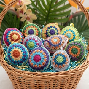 May include: A wicker basket containing a collection of colourful, crocheted Easter egg decorations. The eggs display detailed patterns in shades of blue, orange, purple, and green. Each egg has a ribbon loop. The basket is lined with blue and gold decorative filler.