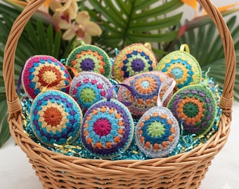 May include: A wicker basket containing a collection of colourful, crocheted Easter egg decorations. The eggs display detailed patterns in shades of blue, orange, purple, and green. Each egg has a ribbon loop. The basket is lined with blue and gold decorative filler.