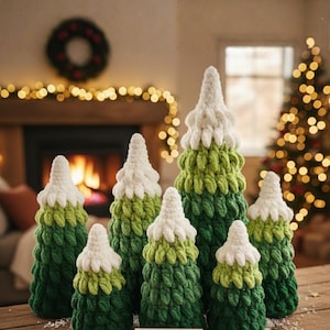 May include: A collection of handmade, ombre-style crochet Christmas trees in varying sizes. The trees feature shades of green and white, with a loopy texture. The scene is set on a wooden surface with a festive background, including a fireplace and Christmas tree.