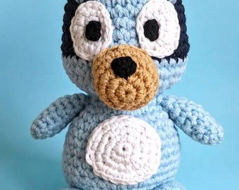 May include: A handmade crochet plush toy shaped like a blue dog with dark blue ears, a tan muzzle, and a black nose. The figure features large white eye patches with black pupils and a circular white patch on the belly, crafted with light blue yarn.