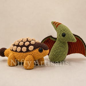 Puede incluir: Dos juguetes de dinosaurio tejidos a ganchillo. Uno es un anquilosaurio de color marrón dorado con cabeza marrón oscura y placas dorsales circulares beige. El otro es un pterodáctilo verde con alas marrón oscuro y naranja, cresta y ojos negros.
