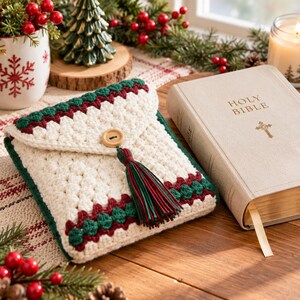 Puede incluir: Una funda de libro tejida a ganchillo de color crema con una solapa triangular, un botón de madera y una borla multicolor en rojo, verde y crema. La funda tiene bordes decorativos en hilo verde oscuro y rojo intenso, junto a una Santa Biblia.
