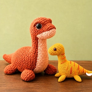 May include: Two crocheted dinosaur toys. The larger dinosaur is rust-orange with a light peach-colored chest and large black eyes. The smaller dinosaur is yellow with orange stripes along its back and sides, featuring small black eyes and a white tail tip.