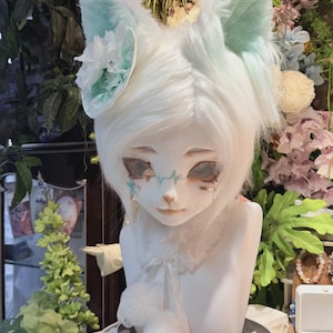 May include: A white cat-eared headpiece with light blue accents. The ears are lined with light blue and adorned with white flowers. The face has detailed makeup, and the neck has a fluffy white collar. The headpiece is displayed on a white mannequin head.