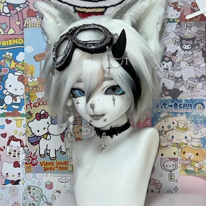 May include: A white mannequin head with a stylized anime aesthetic, featuring white and gray hair styled with cat ears and black accents. It wears goggles, a choker, and cross pendants. The background is decorated with colorful cartoon imagery.