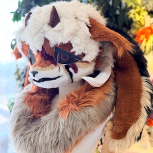 May include: A detailed, furry headpiece with a fox-like design. The headpiece features a white, shaggy top, brown horns, and a mix of orange, brown, and white fur. It has a stylized face with dark accents and a white neck ruff.