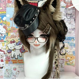May include: A mannequin head with a brown and tan furry wig, cat ears, and a black leather cap with silver chain detailing. The mannequin wears glasses and has a small, open-mouthed expression. The background features Hello Kitty-themed decorations.