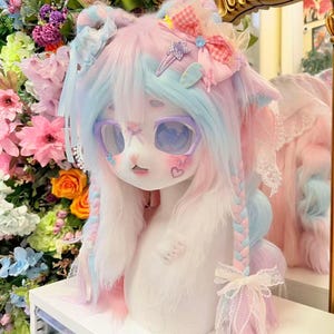 Op de afbeelding: Een pastelkleurige pop met roze en blauw haar, gestyled in vlechten en versierd met strikken en clips. De pop heeft grote, ronde ogen, een bril en een hartvormig detail. De pop staat op een wit oppervlak, met bloemen op de achtergrond.